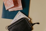 A leather zippered wallet is placed alongside a stack of paper money, including Canadian 50 dollar bills and British 20 pound notes, resting on a blue passport. The wallet is black with visible stitching and a small zipper. The arrangement sits on a light-colored surface with a small coin partially visible at the bottom.