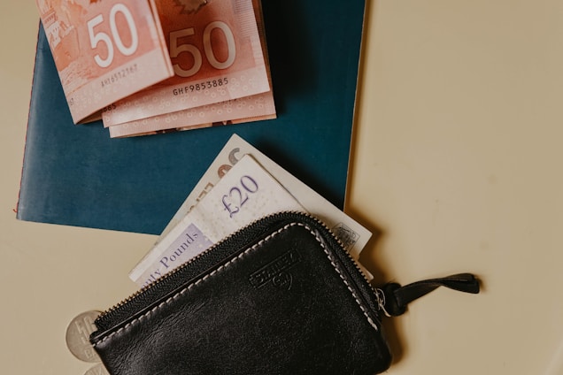 A leather zippered wallet is placed alongside a stack of paper money, including Canadian 50 dollar bills and British 20 pound notes, resting on a blue passport. The wallet is black with visible stitching and a small zipper. The arrangement sits on a light-colored surface with a small coin partially visible at the bottom.