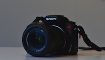 A black Sony DSLR camera with a visible lens and strap, resting on a light surface. The camera has a red alpha symbol and details indicating 10.2 megapixels. The lens has text around the rim, and the overall appearance is clean and professional.