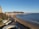 A coastal town with a sandy beach and a promenade next to a wide bay. Buildings, likely part of a town, are situated on the hillside leading down to the water. The scene includes a lighthouse toward the end of a pier, a bus and several cars on a road running parallel to the coast, and a few people walking along the beach. The land extends into the sea forming a headland.