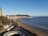 A coastal town with a sandy beach and a promenade next to a wide bay. Buildings, likely part of a town, are situated on the hillside leading down to the water. The scene includes a lighthouse toward the end of a pier, a bus and several cars on a road running parallel to the coast, and a few people walking along the beach. The land extends into the sea forming a headland.