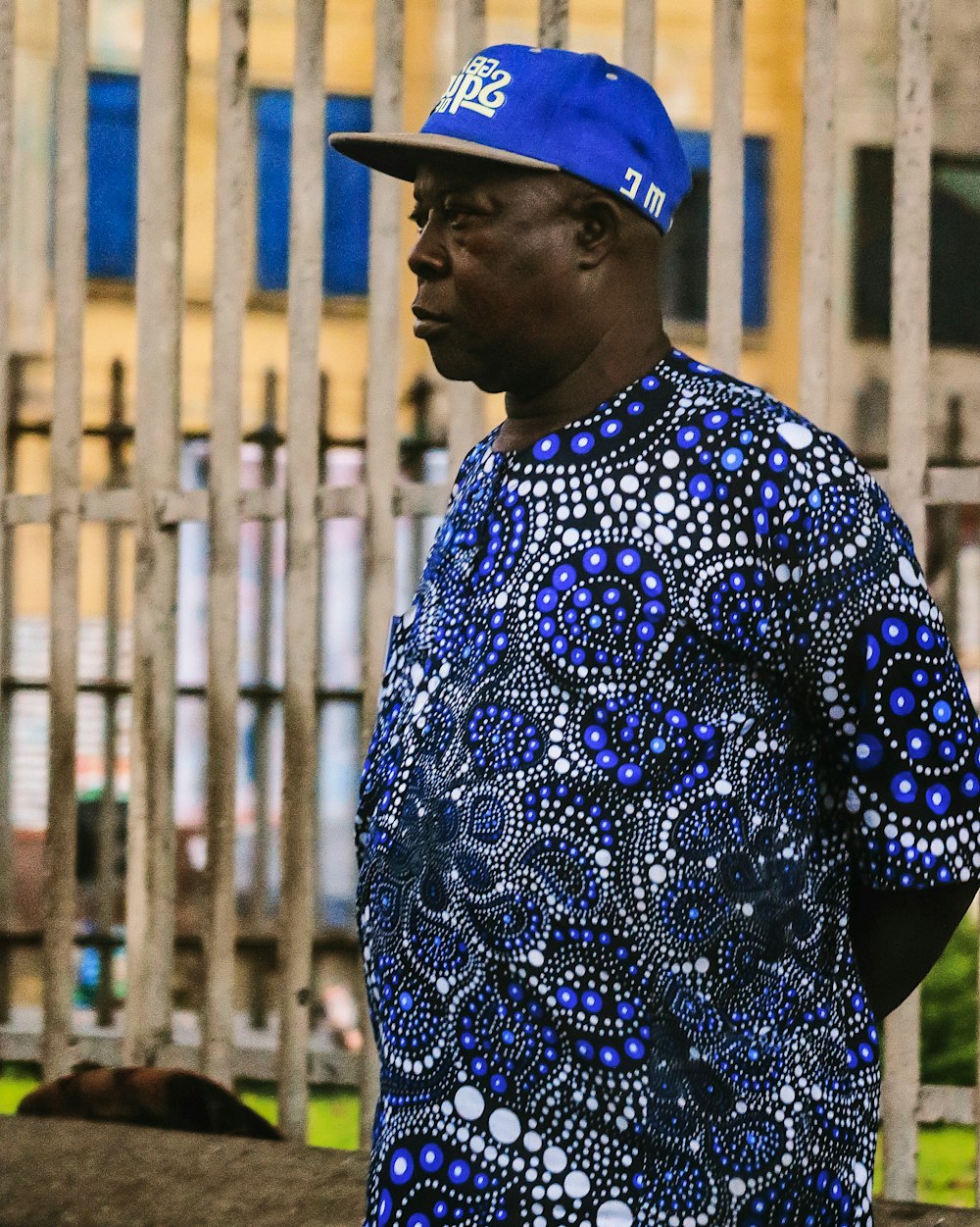 man in blue and white floral shirt wearing blue cap standing near brown metal fence during