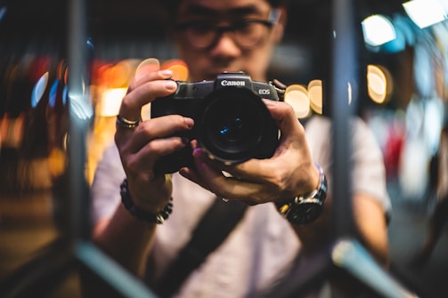 A person holding a Canon EOS camera with both hands, focusing intently on photographing something. The background is a blur of colorful bokeh lights, suggesting a vibrant urban setting at night. The image captures a moment of concentration and creativity.