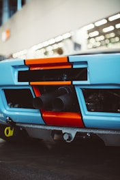 The image features the rear view of a car with a distinctive blue and orange color scheme. Visible are the exhaust pipes protruding from the back, with bold geometric shapes accentuating the design. The background is blurred, suggesting an indoor setting with overhead lighting.