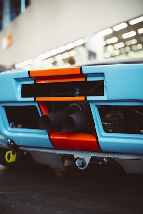 The image features the rear view of a car with a distinctive blue and orange color scheme. Visible are the exhaust pipes protruding from the back, with bold geometric shapes accentuating the design. The background is blurred, suggesting an indoor setting with overhead lighting.