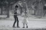 A young boy and a girl are walking hand in hand on a dirt path surrounded by trees. The boy, wearing a striped shirt and jeans, appears to be gently leading the younger girl dressed in a light, fluffy dress. The background features a blurred view of natural greenery, conveying a serene outdoor setting.