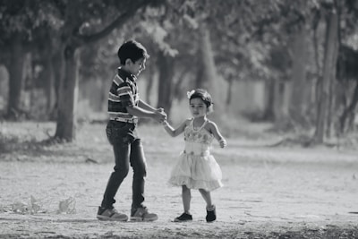 A young boy and a girl are walking hand in hand on a dirt path surrounded by trees. The boy, wearing a striped shirt and jeans, appears to be gently leading the younger girl dressed in a light, fluffy dress. The background features a blurred view of natural greenery, conveying a serene outdoor setting.