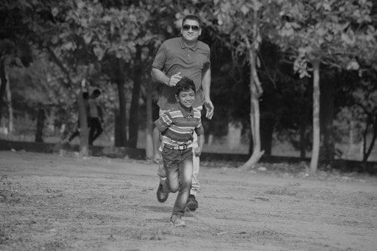 A black and white photo depicting a man and a child running joyfully together in a park. They appear to be on a dirt path with trees providing a natural backdrop, suggesting a playful and carefree atmosphere. The man is wearing sunglasses and casually dressed, while the child is in a striped shirt and jeans.