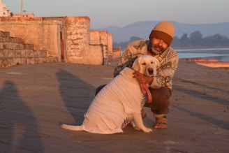 A person wearing a beanie and patterned sweater is kneeling and affectionately hugging a dog dressed in a white shirt. They are on a stone pathway with ancient stone structures and steps in the background, situated beside a body of water. The early morning or late afternoon light casts long shadows, and distant hills are visible under a clear blue sky.