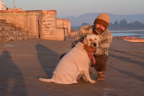 A person wearing a beanie and patterned sweater is kneeling and affectionately hugging a dog dressed in a white shirt. They are on a stone pathway with ancient stone structures and steps in the background, situated beside a body of water. The early morning or late afternoon light casts long shadows, and distant hills are visible under a clear blue sky.