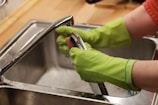 A person wearing bright green rubber gloves is washing a kitchen tap over a stainless steel sink. The setting appears to be a domestic kitchen with a wooden countertop.