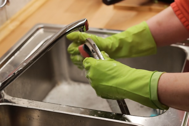A person wearing bright green rubber gloves is washing a kitchen tap over a stainless steel sink. The setting appears to be a domestic kitchen with a wooden countertop.