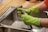 A person wearing bright green rubber gloves is washing a kitchen tap over a stainless steel sink. The setting appears to be a domestic kitchen with a wooden countertop.