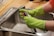 A person wearing bright green rubber gloves is washing a kitchen tap over a stainless steel sink. The setting appears to be a domestic kitchen with a wooden countertop.