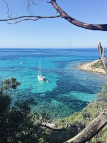 A tranquil turquoise sea framed by rocky coastline and trees with a small sailboat gently floating in the clear waters.