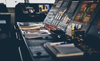 A control room with a large control panel featuring numerous buttons, switches, and screens. The panel is covered with papers and office supplies such as notebooks and highlighters. Displays on the control panel show various indicators and graphical information.