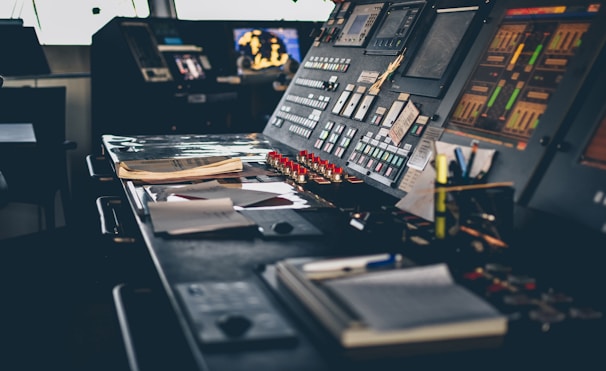 A control room with a large control panel featuring numerous buttons, switches, and screens. The panel is covered with papers and office supplies such as notebooks and highlighters. Displays on the control panel show various indicators and graphical information.