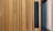 Wooden slats arranged vertically with alternating narrow gaps. The slats have a warm brown tone, and there is a dark blue vertical panel integrated into the arrangement. On the right, glass and metal features are visible, possibly indicating a window or partition.
