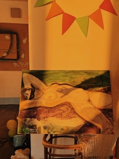 A painting depicting a reclining figure with abstract and impressionistic features, set against a backdrop of rich earthy tones. The painting is displayed on a wooden stool under warm, golden lighting. In the background, there are colorful triangular flags hanging on the wall, creating a festive atmosphere.
