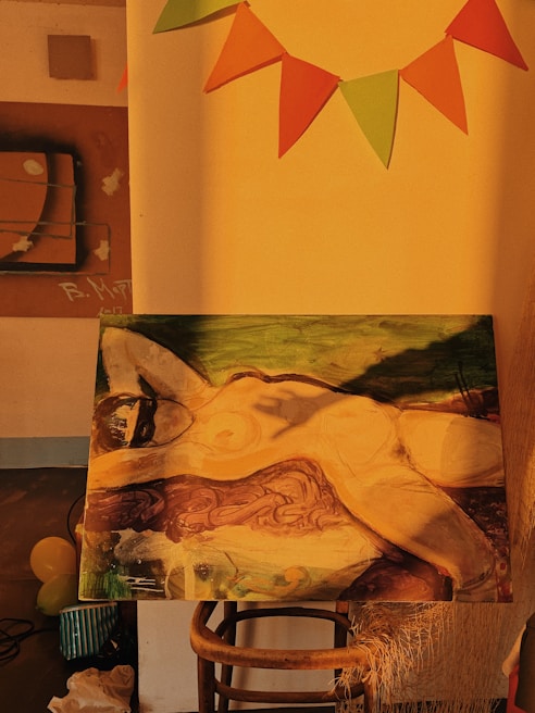 A painting depicting a reclining figure with abstract and impressionistic features, set against a backdrop of rich earthy tones. The painting is displayed on a wooden stool under warm, golden lighting. In the background, there are colorful triangular flags hanging on the wall, creating a festive atmosphere.