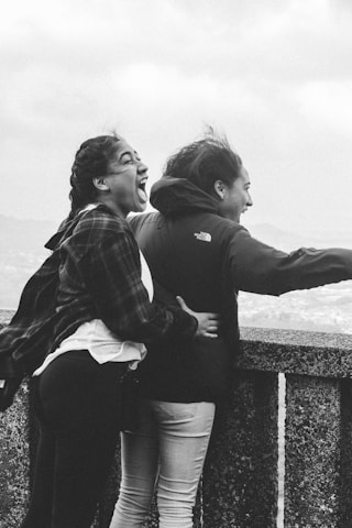 Two individuals are captured in a candid moment of joy, standing at a vantage point with an expansive view behind them. The wind appears to be blowing, adding a sense of dynamic movement to their hair and clothing. One person is laughing exuberantly, with an expression of happiness and excitement.