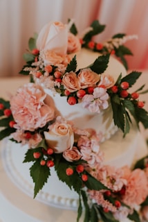 A beautifully decorated cake adorned with pink roses, peonies, and small red berries. The cake has lush green leaves interspersed among the flowers, enhancing its elegant and fresh appearance. The soft, pastel colors and delicate textures of the flowers give it a romantic and sophisticated look.