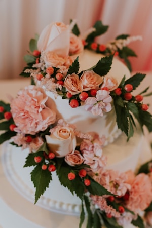 A beautifully decorated cake adorned with pink roses, peonies, and small red berries. The cake has lush green leaves interspersed among the flowers, enhancing its elegant and fresh appearance. The soft, pastel colors and delicate textures of the flowers give it a romantic and sophisticated look.