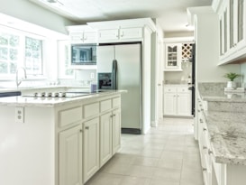 A spacious, modern kitchen featuring white cabinetry and light grey granite countertops. There is a large stainless steel refrigerator, a built-in microwave above the counters, a stovetop on the island, and a wine rack in the corner. Large windows fill the space with natural light, and a small potted plant decorates the countertop.