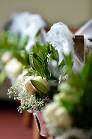 A close-up of floral arrangements featuring white roses, baby's breath, and lush green leaves, adorned with delicate white tulle bows on what appears to be a church pew.