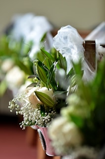 A close-up of floral arrangements featuring white roses, baby's breath, and lush green leaves, adorned with delicate white tulle bows on what appears to be a church pew.