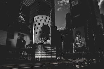 A dark, monochromatic cityscape featuring large billboard advertisements on the sides of tall buildings. The image conveys a sense of urban life with the focus on modern, graphic advertising, including a prominent light-adorned column with a silhouette of a person's profile and the text 'REWRITE TOMORROW'. Another advertisement features a person holding something to eat with the text 'MEET LCT' below.