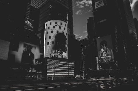 A dark, monochromatic cityscape featuring large billboard advertisements on the sides of tall buildings. The image conveys a sense of urban life with the focus on modern, graphic advertising, including a prominent light-adorned column with a silhouette of a person's profile and the text 'REWRITE TOMORROW'. Another advertisement features a person holding something to eat with the text 'MEET LCT' below.