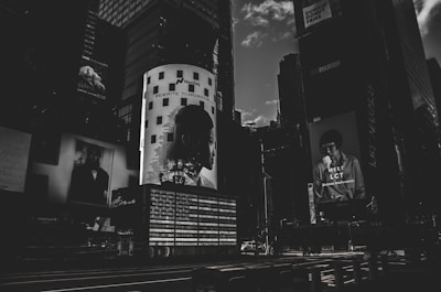 A dark, monochromatic cityscape featuring large billboard advertisements on the sides of tall buildings. The image conveys a sense of urban life with the focus on modern, graphic advertising, including a prominent light-adorned column with a silhouette of a person's profile and the text 'REWRITE TOMORROW'. Another advertisement features a person holding something to eat with the text 'MEET LCT' below.