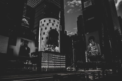 A dark, monochromatic cityscape featuring large billboard advertisements on the sides of tall buildings. The image conveys a sense of urban life with the focus on modern, graphic advertising, including a prominent light-adorned column with a silhouette of a person's profile and the text 'REWRITE TOMORROW'. Another advertisement features a person holding something to eat with the text 'MEET LCT' below.
