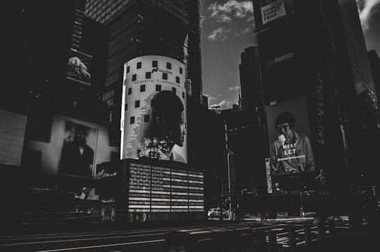 A dark, monochromatic cityscape featuring large billboard advertisements on the sides of tall buildings. The image conveys a sense of urban life with the focus on modern, graphic advertising, including a prominent light-adorned column with a silhouette of a person's profile and the text 'REWRITE TOMORROW'. Another advertisement features a person holding something to eat with the text 'MEET LCT' below.