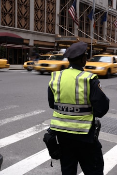 A police officer wearing a neon yellow and black NYPD vest stands on a city street overseeing the busy traffic. Several yellow taxis blur past on the road, while the backdrop includes a building with large windows and flags mounted above an entrance.