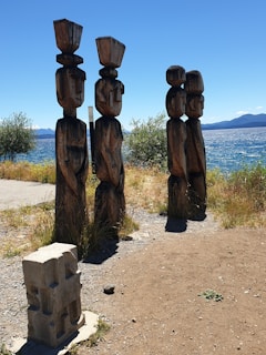 A group of four carved wooden totem poles stands on a dirt path beside a body of water. The poles have distinct geometric shapes and are positioned against a backdrop of trees and mountains under a clear blue sky. In the foreground, there is a small stone sculpture with defined edges and surfaces.