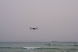 A drone hovers over a calm sea with an overcast sky. The horizon is visible, and small waves are gently breaking in the foreground. Two small islands can be seen in the distance.