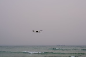 A drone hovers over a calm sea with an overcast sky. The horizon is visible, and small waves are gently breaking in the foreground. Two small islands can be seen in the distance.