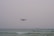 A drone hovers over a calm sea with an overcast sky. The horizon is visible, and small waves are gently breaking in the foreground. Two small islands can be seen in the distance.