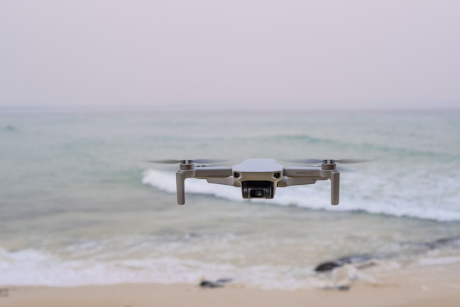 A drone hovers in mid-air above a tranquil beach setting, with gentle waves rolling towards the shore. The ocean extends into the horizon under a pale, serene sky. The drone is centrally positioned, capturing an expansive view.