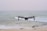 A drone hovers in mid-air above a tranquil beach setting, with gentle waves rolling towards the shore. The ocean extends into the horizon under a pale, serene sky. The drone is centrally positioned, capturing an expansive view.