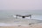 A drone hovers in mid-air above a tranquil beach setting, with gentle waves rolling towards the shore. The ocean extends into the horizon under a pale, serene sky. The drone is centrally positioned, capturing an expansive view.