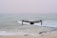 A drone hovers in mid-air above a tranquil beach setting, with gentle waves rolling towards the shore. The ocean extends into the horizon under a pale, serene sky. The drone is centrally positioned, capturing an expansive view.