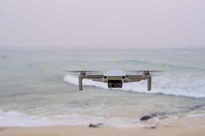 A drone hovers in mid-air above a tranquil beach setting, with gentle waves rolling towards the shore. The ocean extends into the horizon under a pale, serene sky. The drone is centrally positioned, capturing an expansive view.
