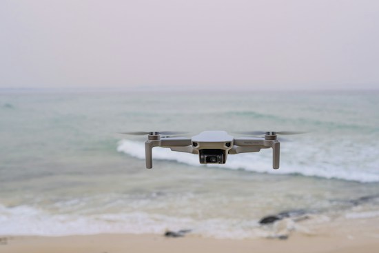 A drone hovers in mid-air above a tranquil beach setting, with gentle waves rolling towards the shore. The ocean extends into the horizon under a pale, serene sky. The drone is centrally positioned, capturing an expansive view.