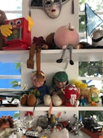A collection of toys and figurines is displayed on two shelves. The upper shelf features a colorful mask, a red toy with yellow arms, and a large soft toy shaped like a pink face. There's also a blue flag in the background. On the lower shelf, several figurines, including cartoon characters, a red M&M's character, and a miniature airplane, are arranged alongside a small disco ball and a model of a cruise ship.