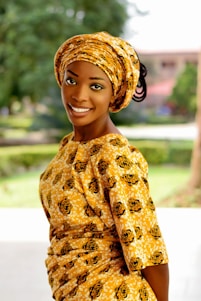A woman is smiling while wearing a traditional African outfit. The fabric has a yellow pattern with black details. She is standing outdoors with green foliage in the background, giving a relaxed and natural setting.