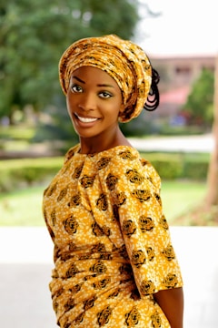 A woman is smiling while wearing a traditional African outfit. The fabric has a yellow pattern with black details. She is standing outdoors with green foliage in the background, giving a relaxed and natural setting.