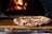 A wood-fired pizza with a charred crust sits on a rustic wooden board. The pizza is topped with melted cheese, tomato sauce, and fresh basil. In the background, a fire is burning in a stone oven, casting a warm glow.